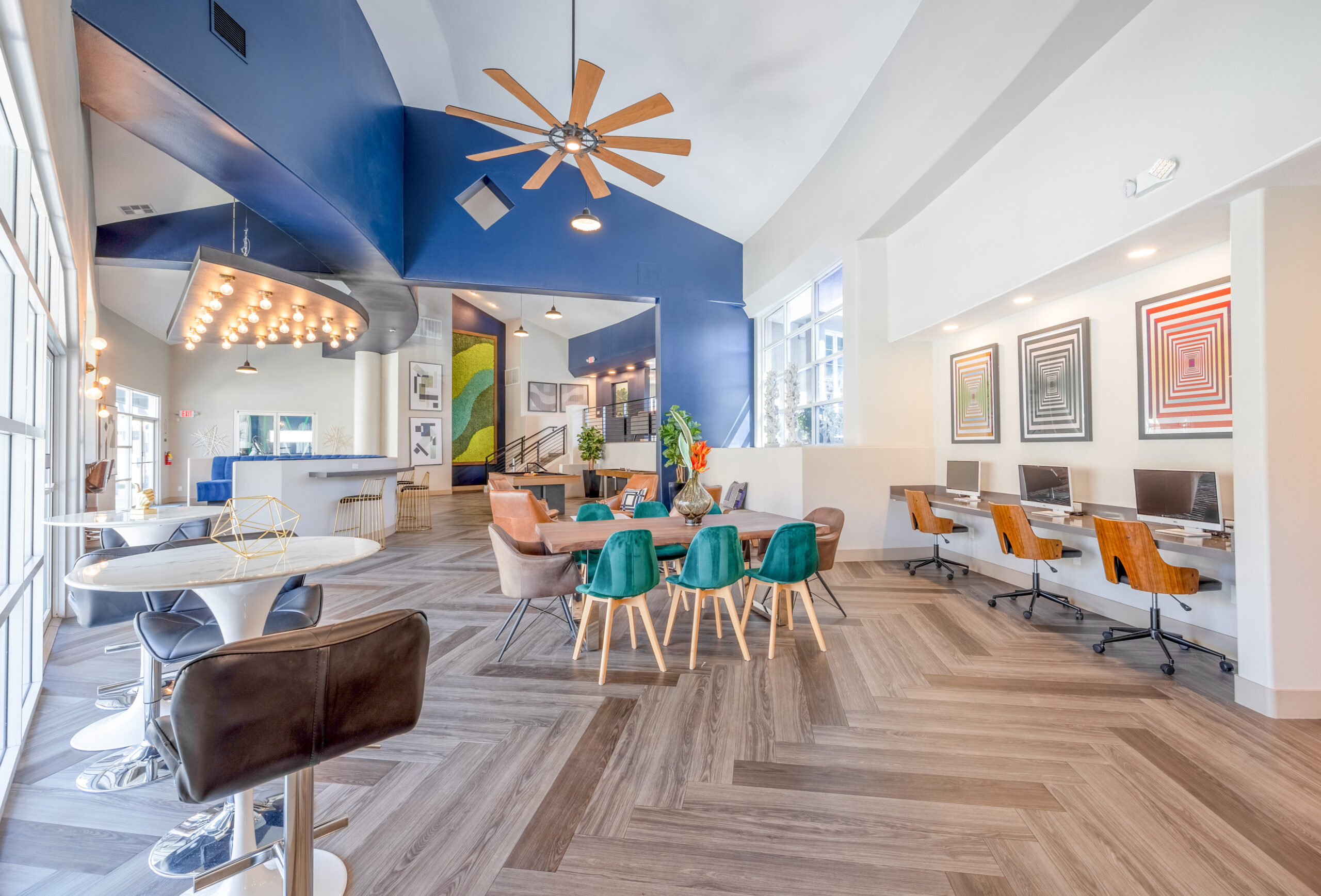 Inside the resident clubhouse is an open space for residents to gather or work from home. A large table with blue chairs, individual work desks along one wall, and a table for two next to the large windows.