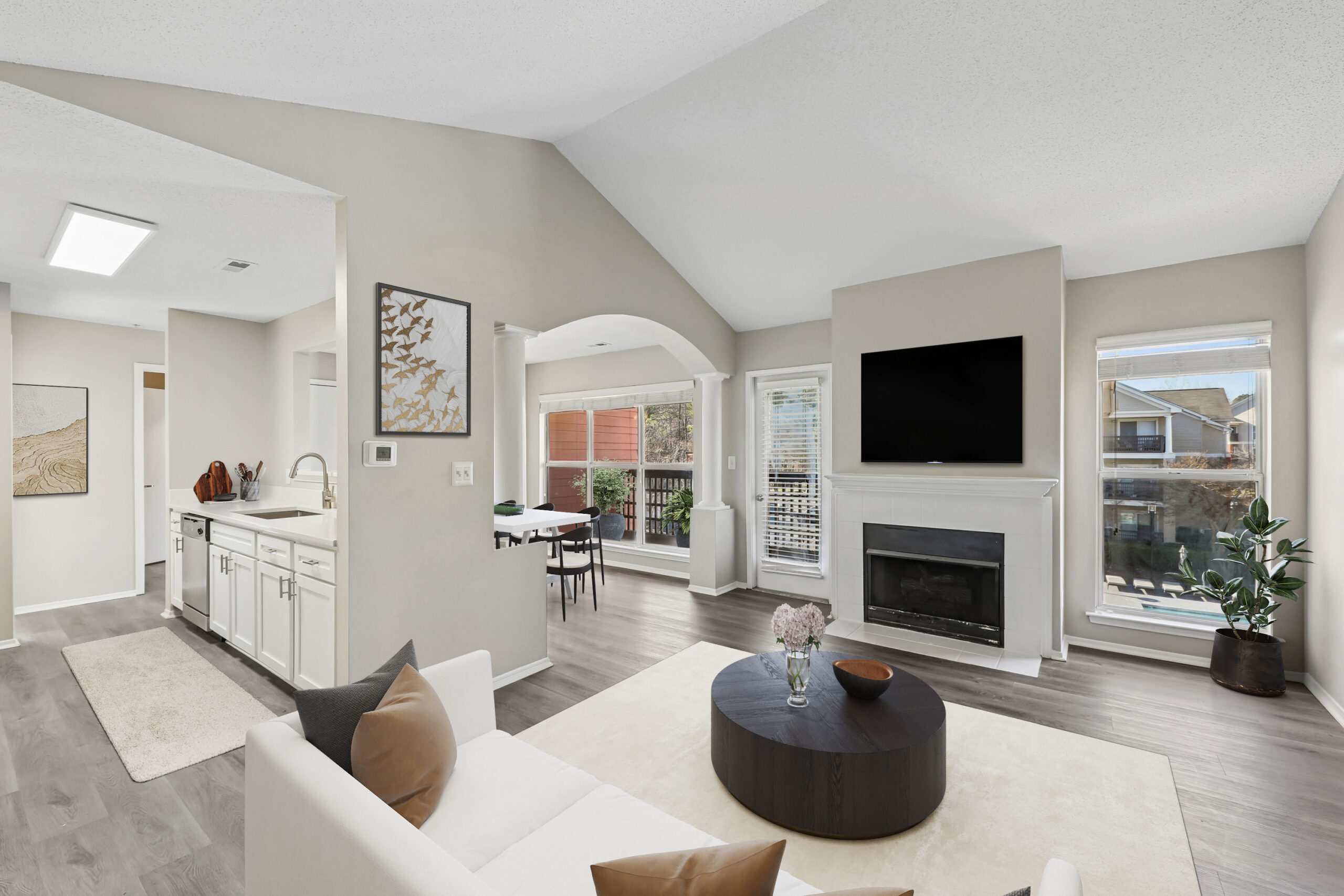 Bright living room with vaulted ceiling, beige walls, and wood floors. A white couch faces a black TV above a fireplace. Large windows reveal outdoor views, enhancing the airy, modern ambiance.