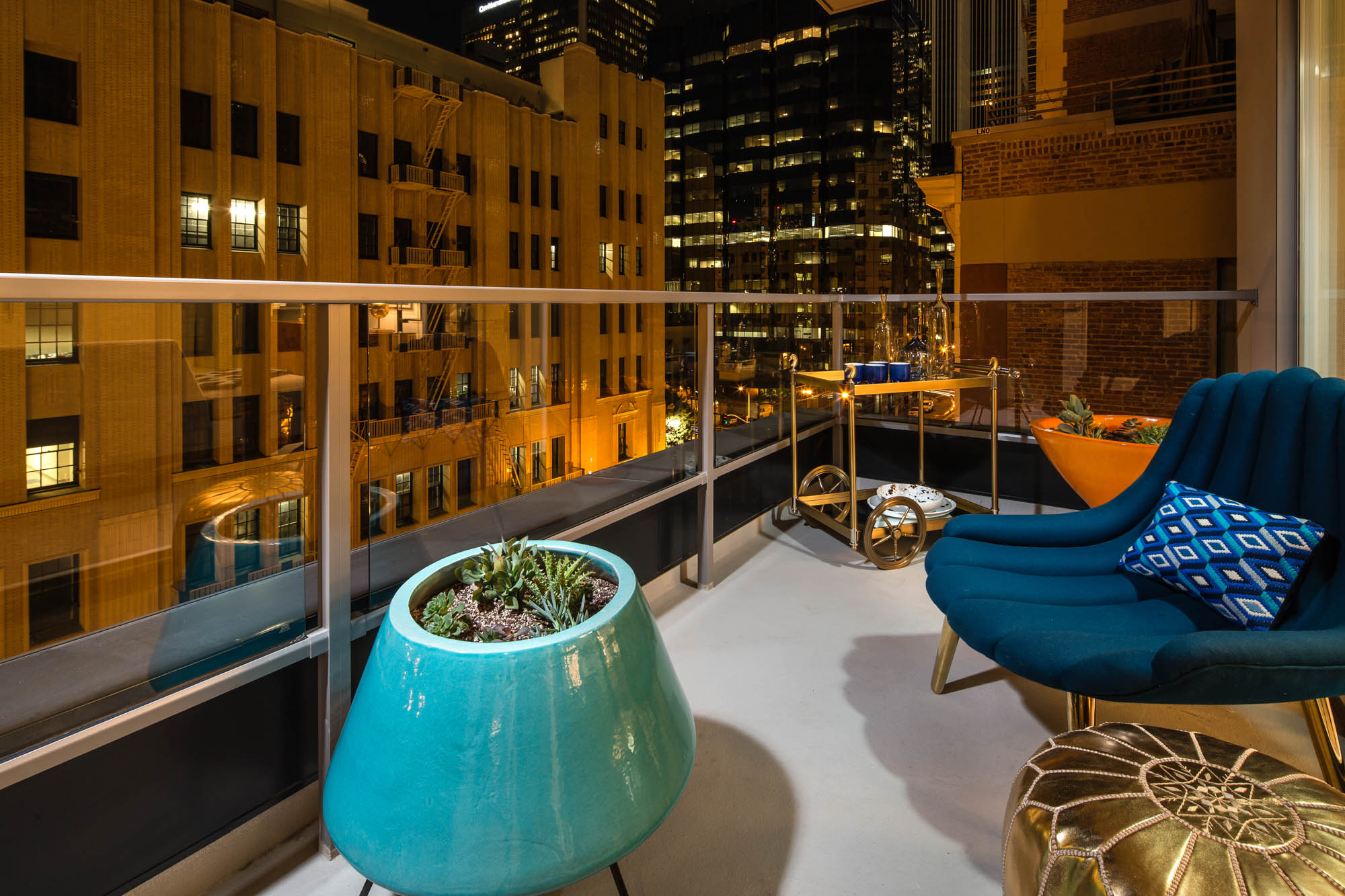 Rooftop balcony at night with cityscape view. Features a teal planter with succulents, a blue chair with a geometric pillow, and a golden ottoman.