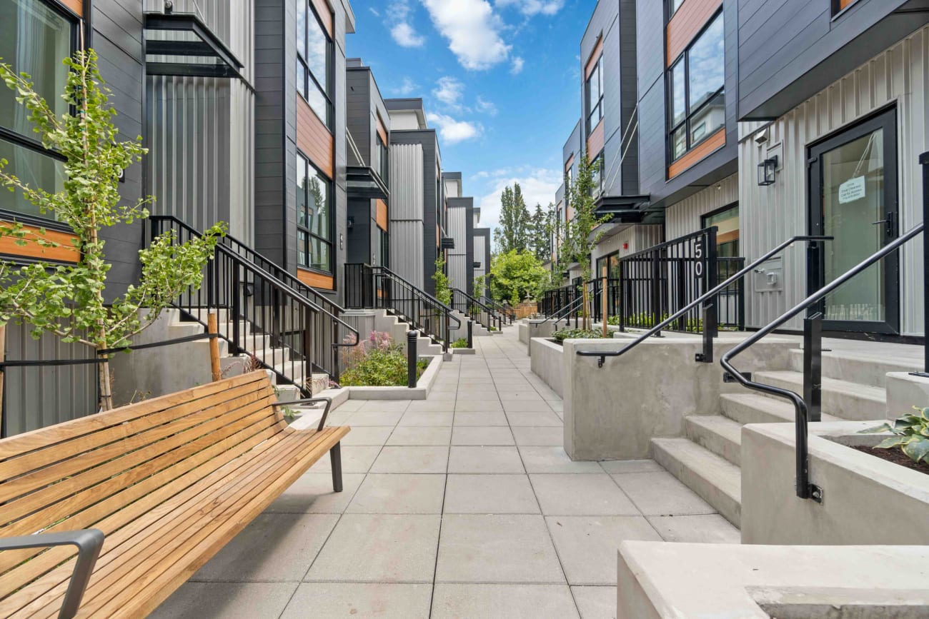 Modern urban courtyard with gray townhouses on either side, featuring large windows and wood accents. A wooden bench and small trees line the walkway. Bright, clear sky.