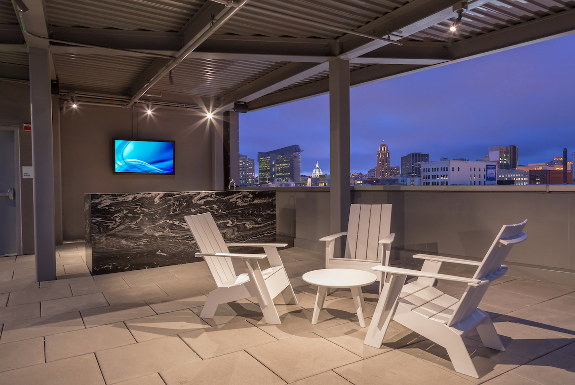 Rooftop terrace at dusk with white Adirondack chairs around a table, a modern black marble counter, city skyline view, and wall-mounted TV.