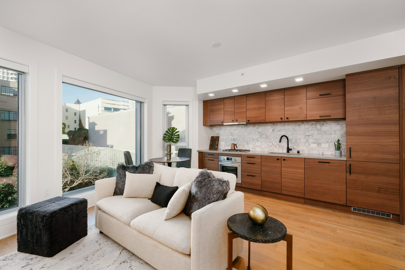 Spacious living room featuring an open-concept kitchen with wooden cabinets, white backsplash, and modern appliances, illuminated by natural light from large windows overlooking a garden.