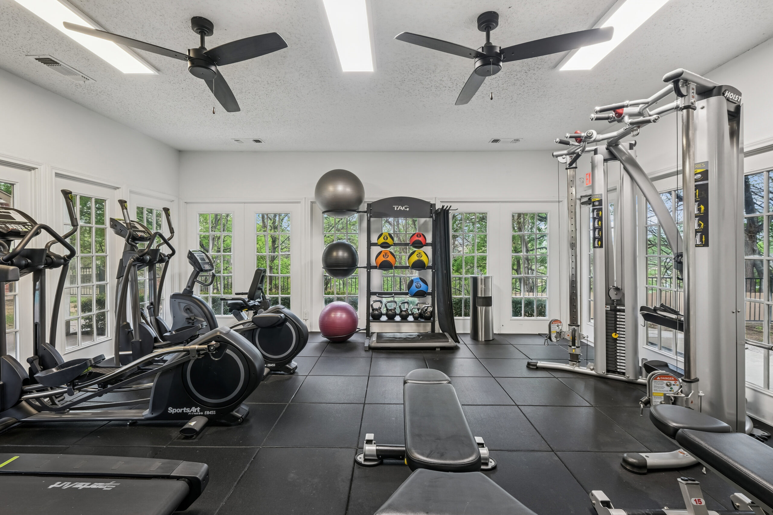 Modern apartment fitness center with cardio machines, free weights, and large windows overlooking green outdoor views on each wall