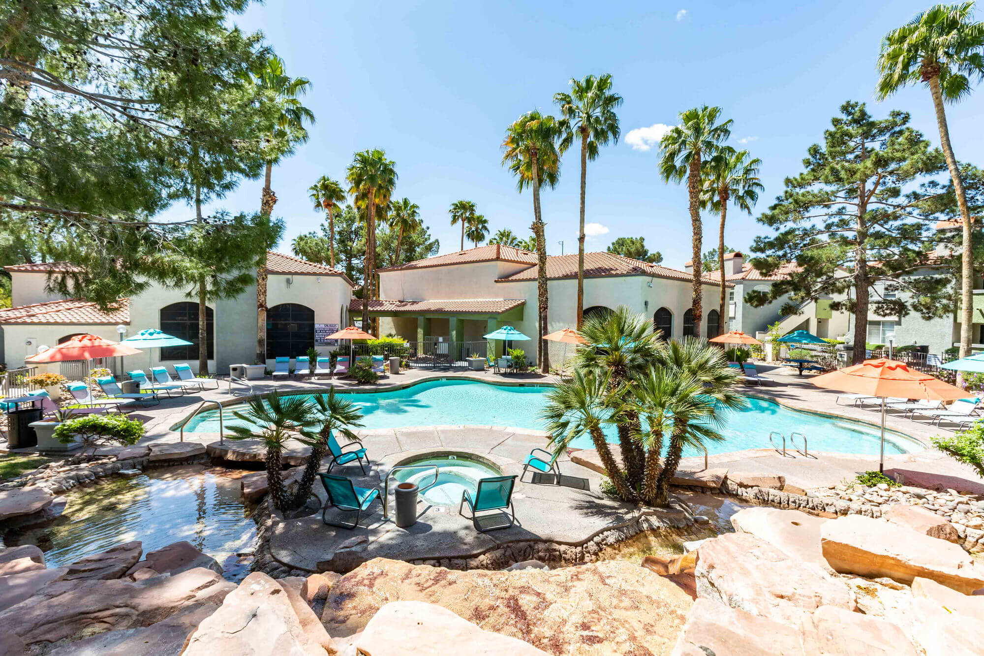 Luxurious outdoor pool area at LyvWell Apartments featuring palm trees, lounge chairs, and shaded seating in a tropical setting.