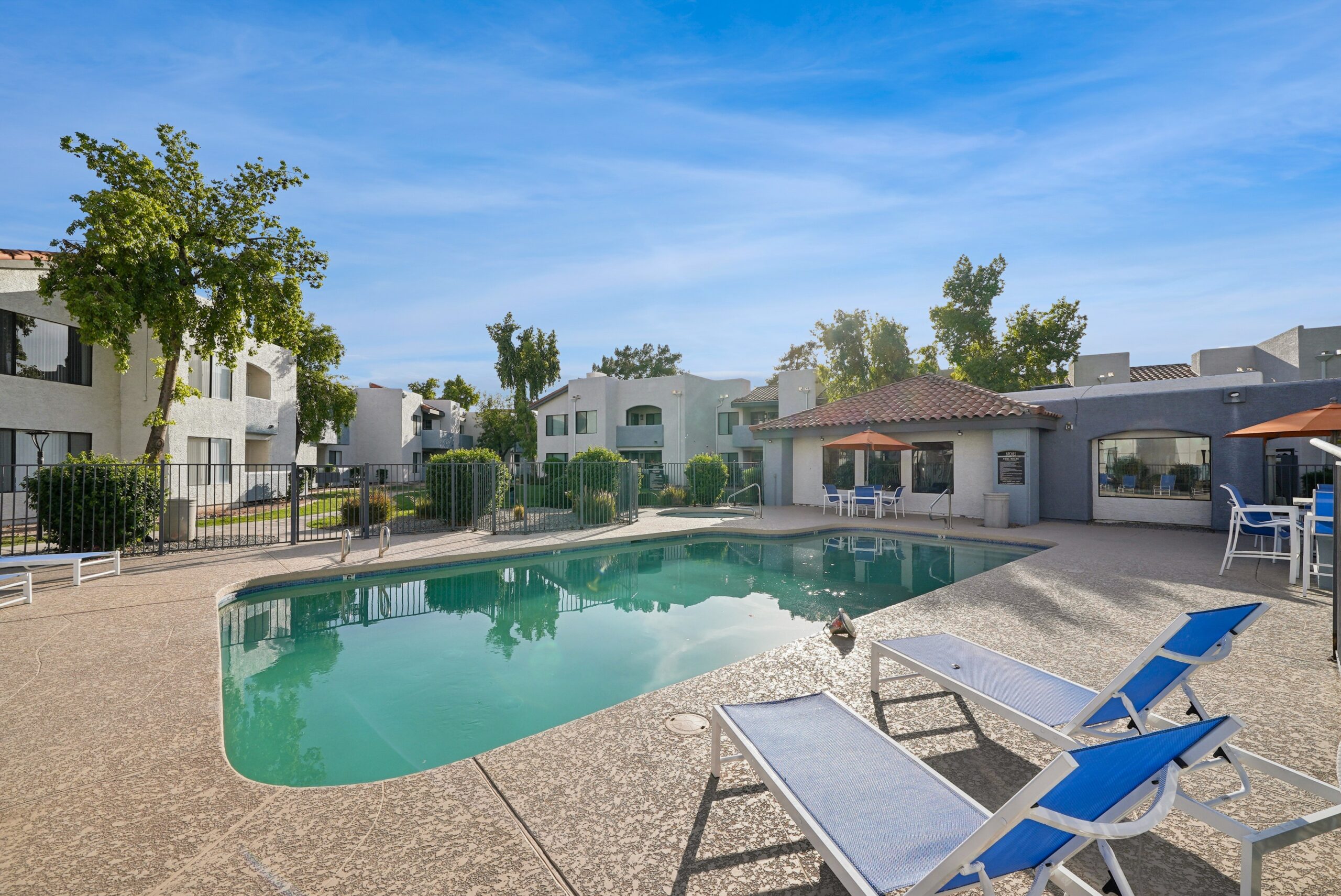 Community swimming pool surrounded by blue lounge chairs, outdoor seating, mature trees, and apartment buildings.